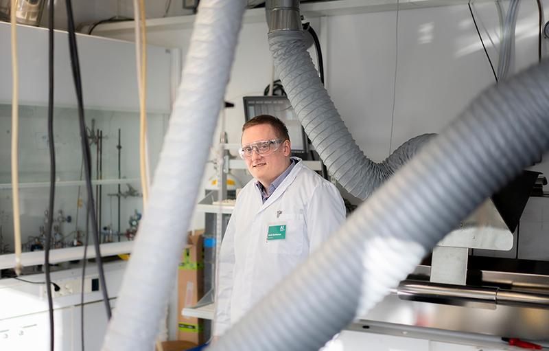 Mies valkoisessa laboratoriotakissa seisoo laboratoriossa putkien takana.