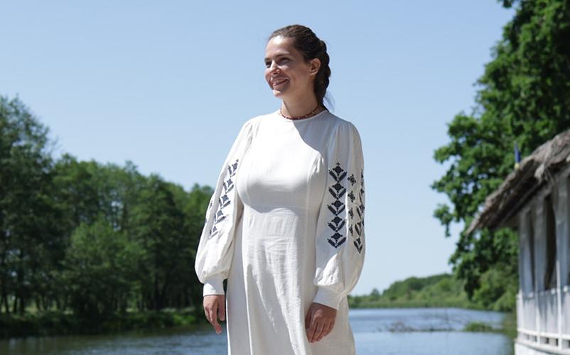 Nainen valkoisessa mekossa seisoo kesäisessä järvimaisemassa.